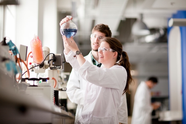 Two students in lab coats examining a round bottom flask with a blue liquid in it