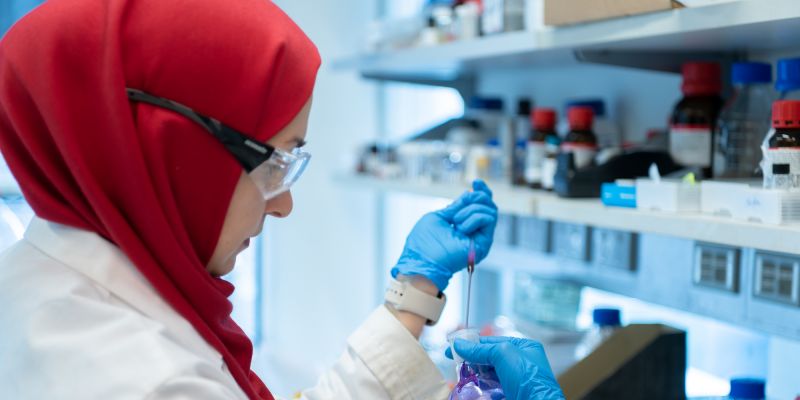 A researcher in PPE and a hijab using a pipette to transfer purple liquid into a round bottom flask